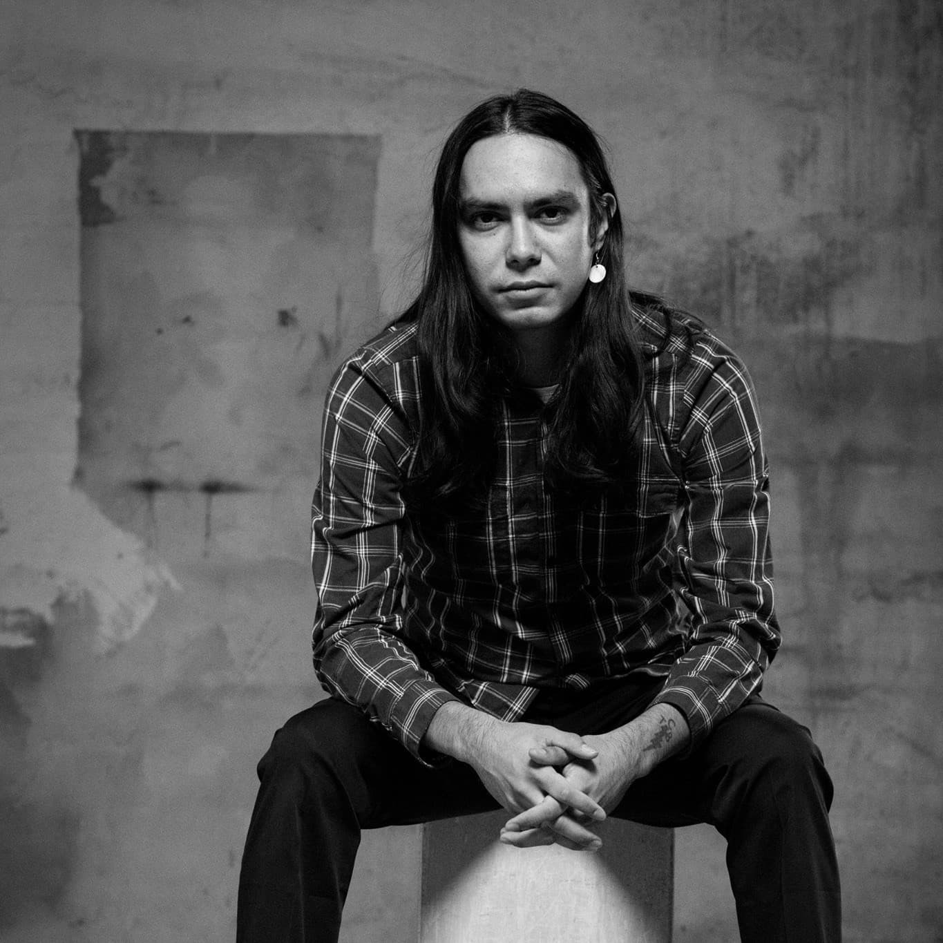 A black -and-white picture of a man named Ishkwaazhe Shane McSauby. He has long, dark, straight hair and is sitting on a wooden crate. He is dressed in a checkered shirt and dark trousers. His hands are clasped in front of him. The background is a textured, somewhat industrial, and his expression is serious and introspective as he directly faces the camera.
