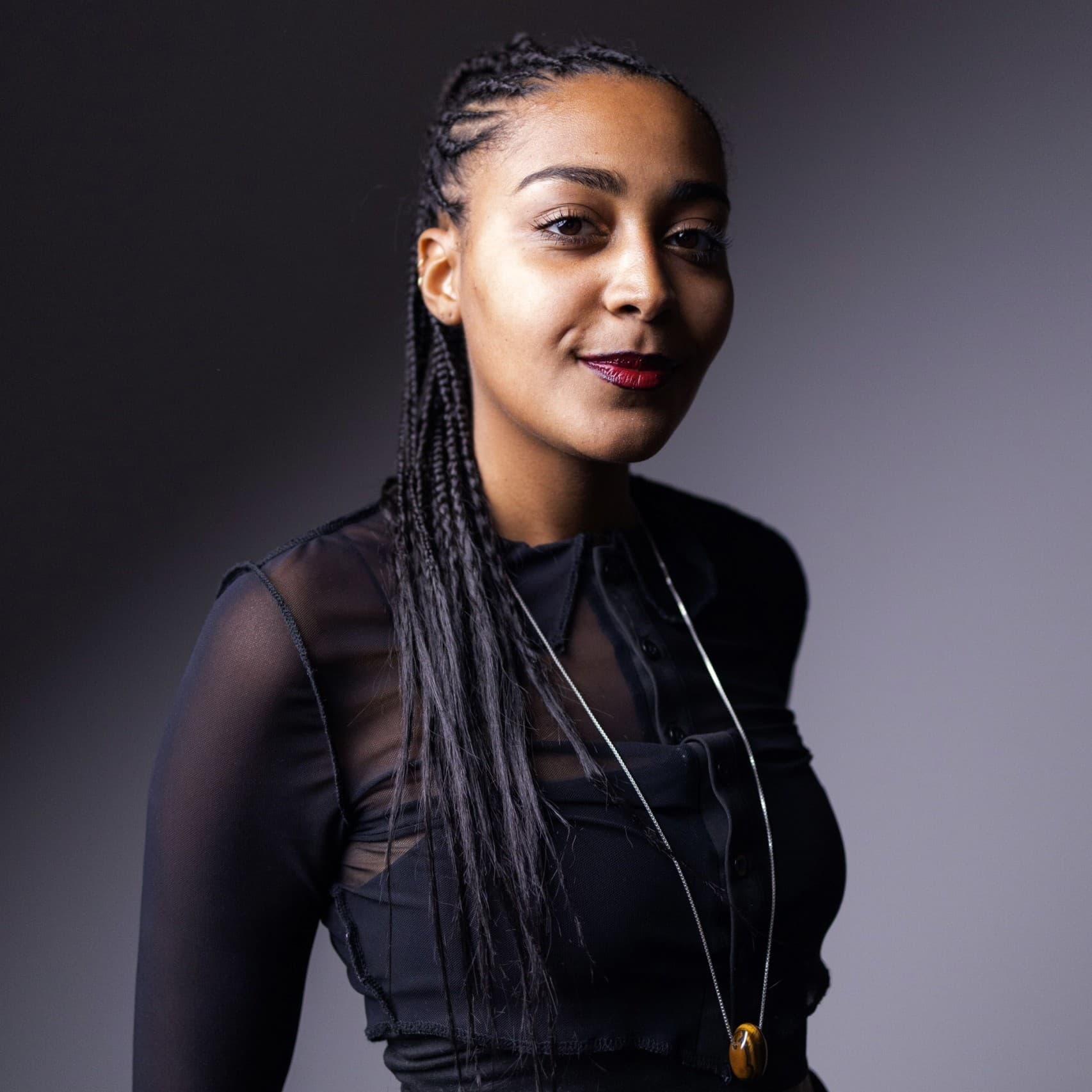 A picture of a woman named Alicia Mendy. She stands confidently against a gradient gray background with a bold red lip. She has long, braided hair that is pulled back neatly. She is wearing a black, long-sleeved, sheer top that reveals another layer underneath paired with a long, thin necklace with a pendant hanging around her neck. She has a slight, poised smile, and her gaze is directed towards the camera.