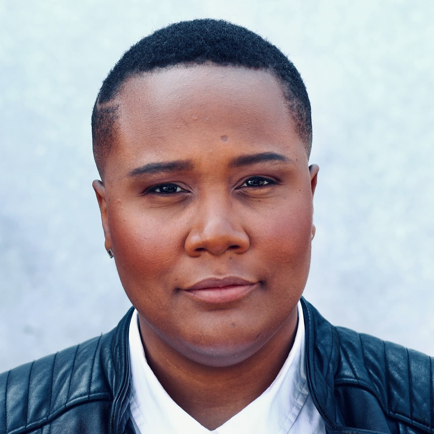 A picture of a woman named Rochee Jeffrey. She is against a soft, out-of-focus background with a light blue and white gradient. She is looking directly into the camera with a calm, composed expression with a slight smile. She has a short, close-cropped haircut and wears a collared shirt under a black leather jacket.