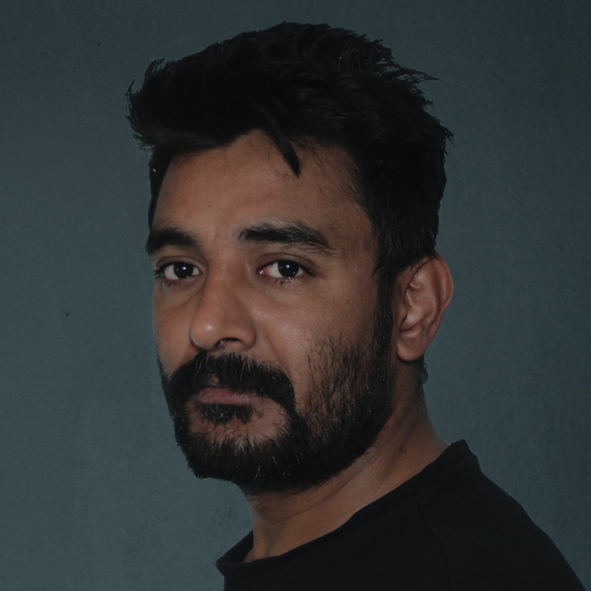 A picture of a man named Niranjan Raj Bhetwal. He is against a plain, dark gray background facing slightly to his left, with his head turned towards the camera. He has short, dark hair that is styled messily. His facial hair consists of a thick, full beard and mustache. He is wearing a black t-shirt and has a calm, serious expression on his face.