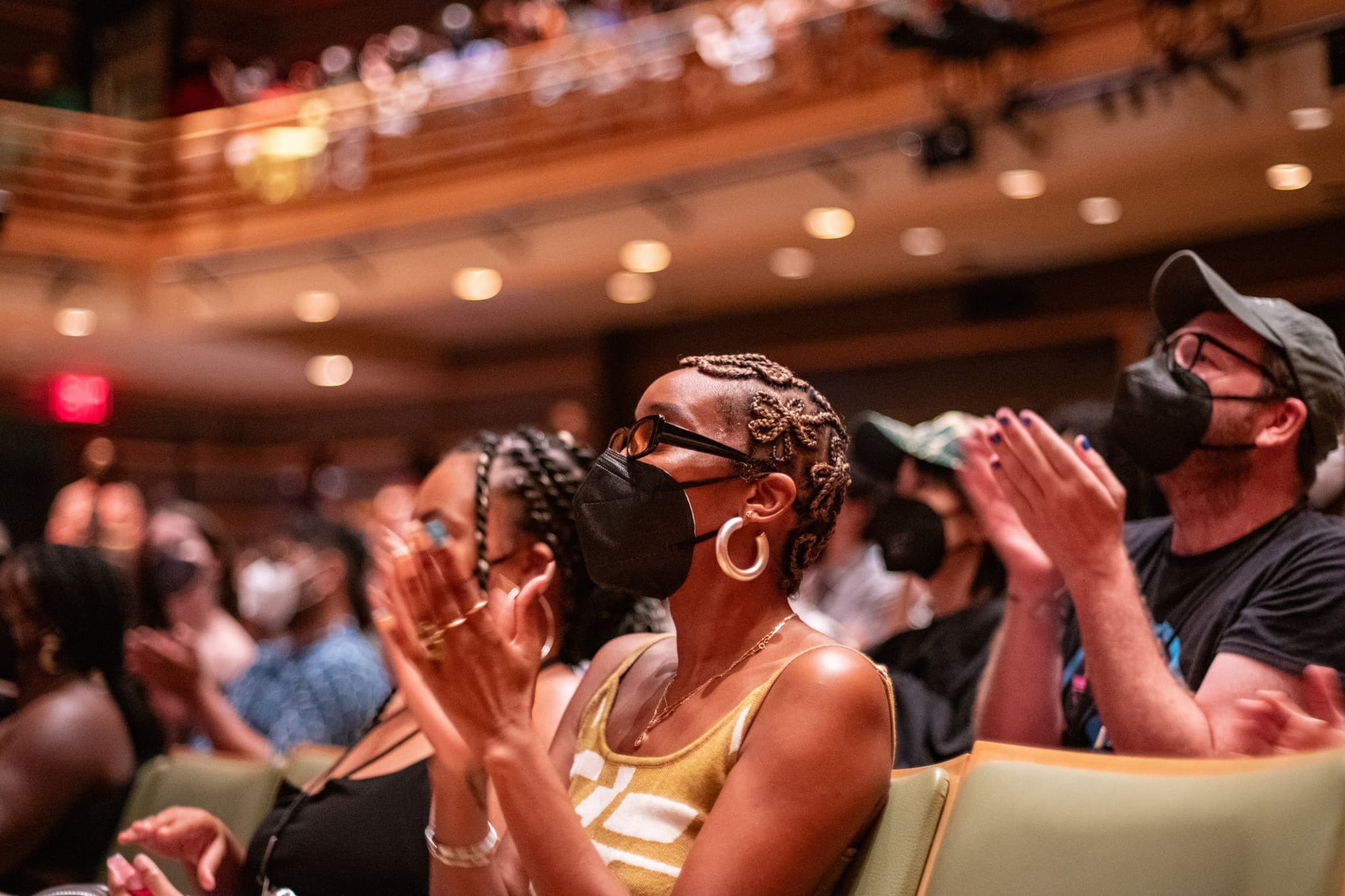 An audience member, wearing a black face mask, claps in the foreground inside a movie theater.