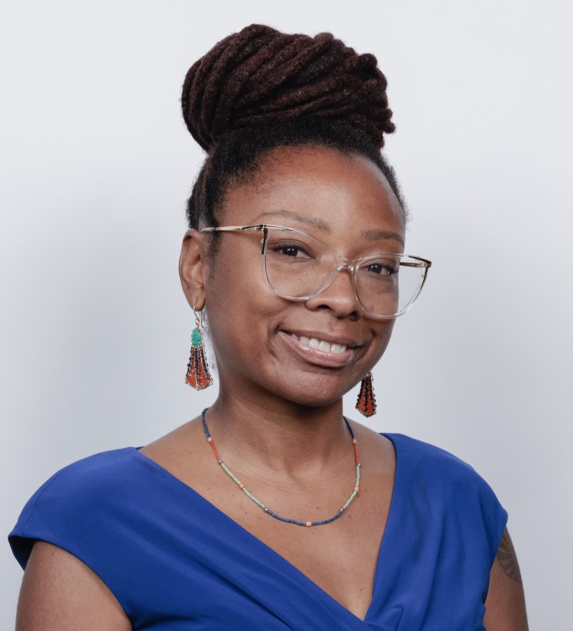 A headshot of Zendra. She is a Black woman, her hair is locked and tied into a bun. She is wearing glasses, earrings, a necklace, and a blue dress.