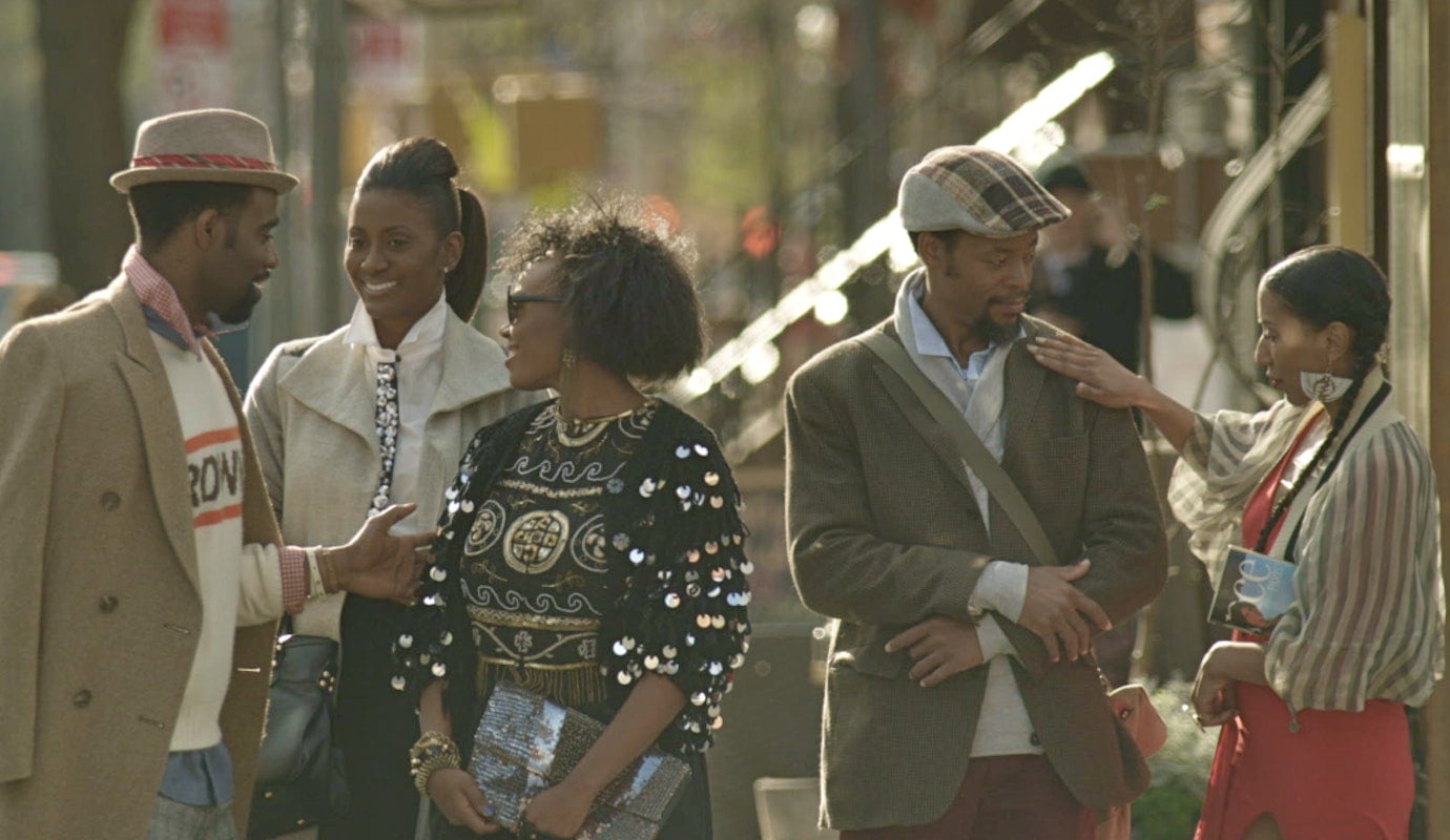 A still from In the Morning shows five Black people, they are are milling around and chatting with one another. Their outfits are 90s style.