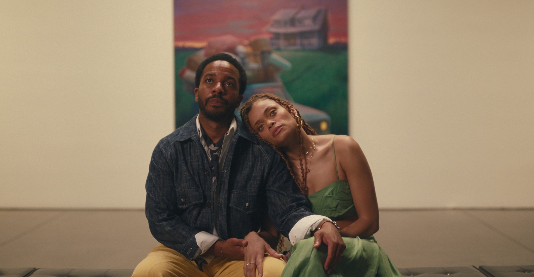 A still from Exhibiting Forgiveness shows a couple sitting in front of a painting in a museum. The woman has her head leaned on the man's shoulder, they are both staring straight ahead with thoughtful expressions.