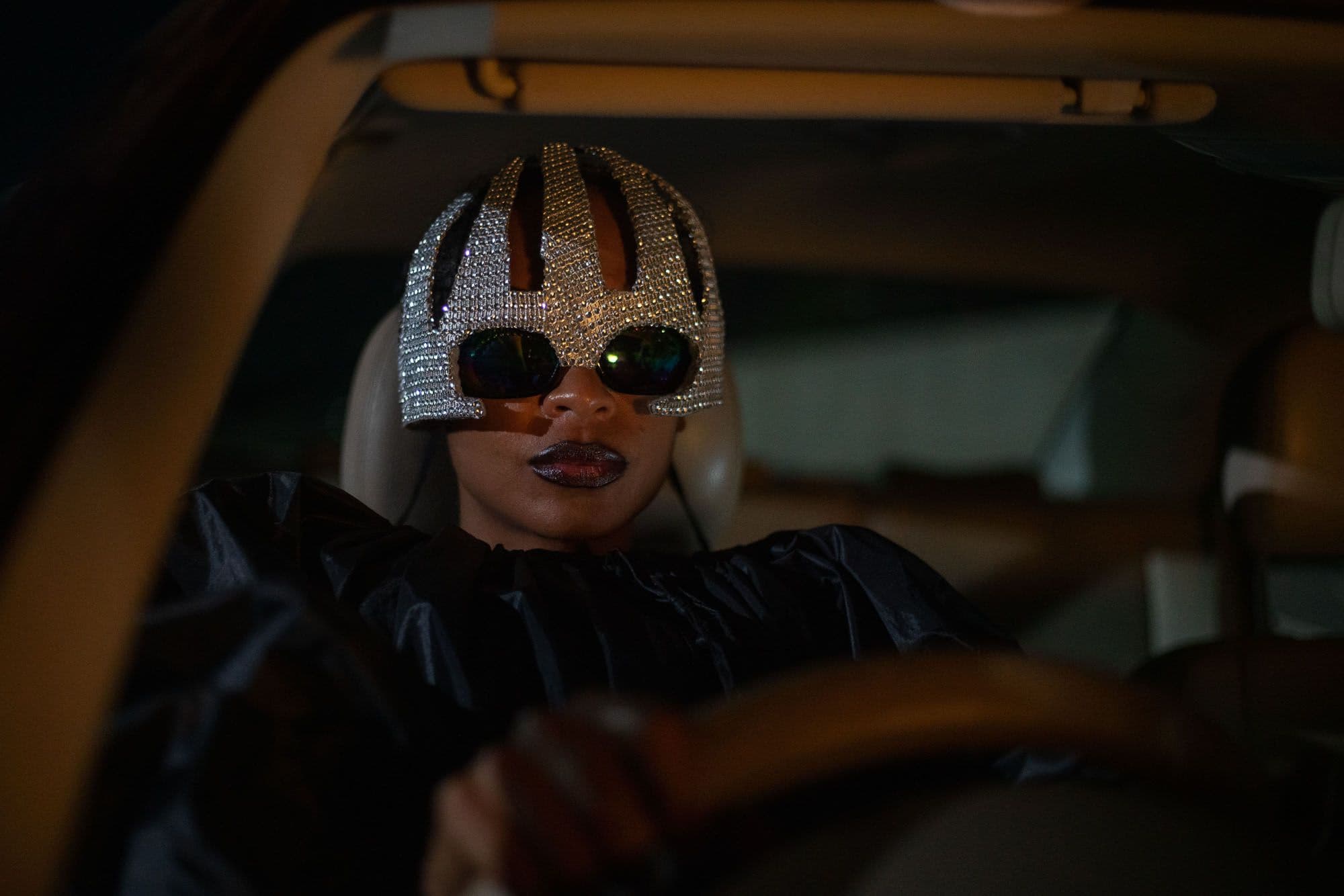 A still from On Becoming A Guinea Fowl shows a woman wearing a futuristic silver cap and sunglasses. She is sitting in a car.