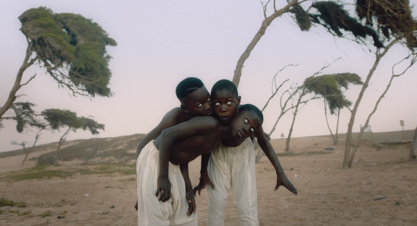 A still from The Dawn shows an arid field. There are three young Black boys standing together, their bodies folded into one another as if they are one. Their eyes are white.