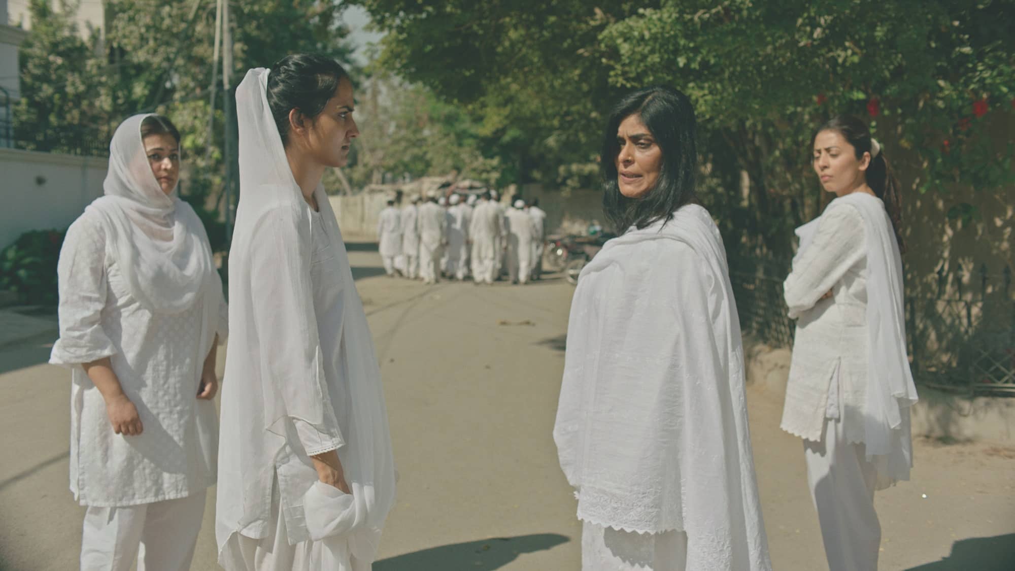 A still image from “The Queen of My Dreams” (2023) showing a young Indian woman in a white saree and head scarf looking at an older Indian woman also wearing a white saree and head scarf. In the middle of the image, another older Indian woman looks at the two women. In the background, a group of men wearing white gowns walk into the distance.