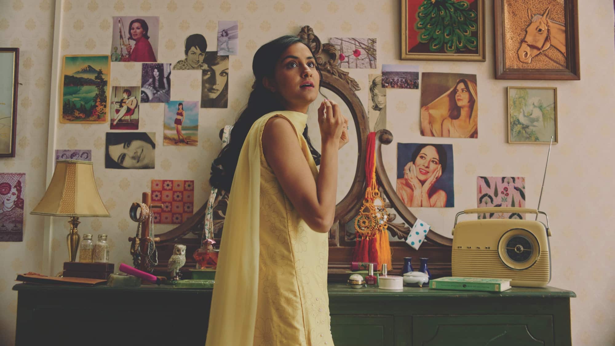 A still image from “The Queen of My Dreams” (2023) showing a young Indian woman with long black hair in a yellow saree in front of her vanity and a wall of posters of Bollywood actresses. She looks away from the vanity to something offscreen.