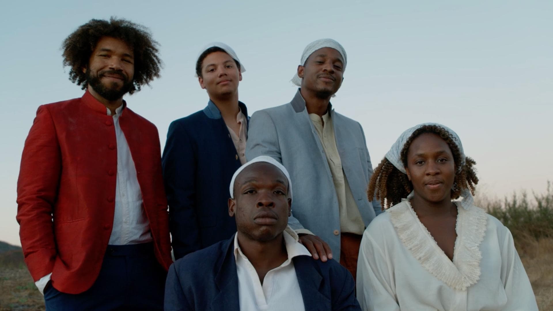 A film still from Boil That Cabbage Down shows a group of Black people posed together in a field with a clear light blue sky. At the center is a man wearing a blue blazer over a white collared shirt. Next to him is a woman wearing a white shirt with a frilly collar. Behind them stand three Black men also in blazers. The man on the left stands out, wearing a red blazer and an afro. He is the only person without a white scarf tied on their head.