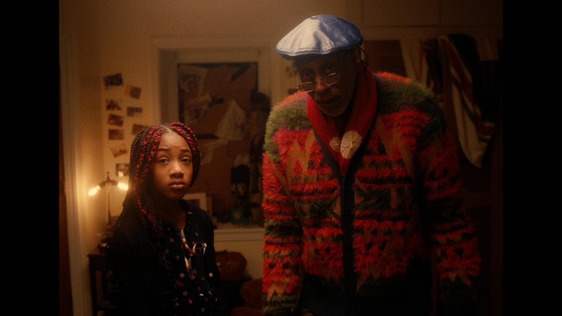 A film still from Della Can Fly shows an older Black man and young Black girl standing next to each other, looking beyond the camera. The man wears a blue beret and a red multicolored fuzzy cardigan. The young girl is wearing a black polka dot top, and her black hair is styled in braids, with the first two on each side being red.