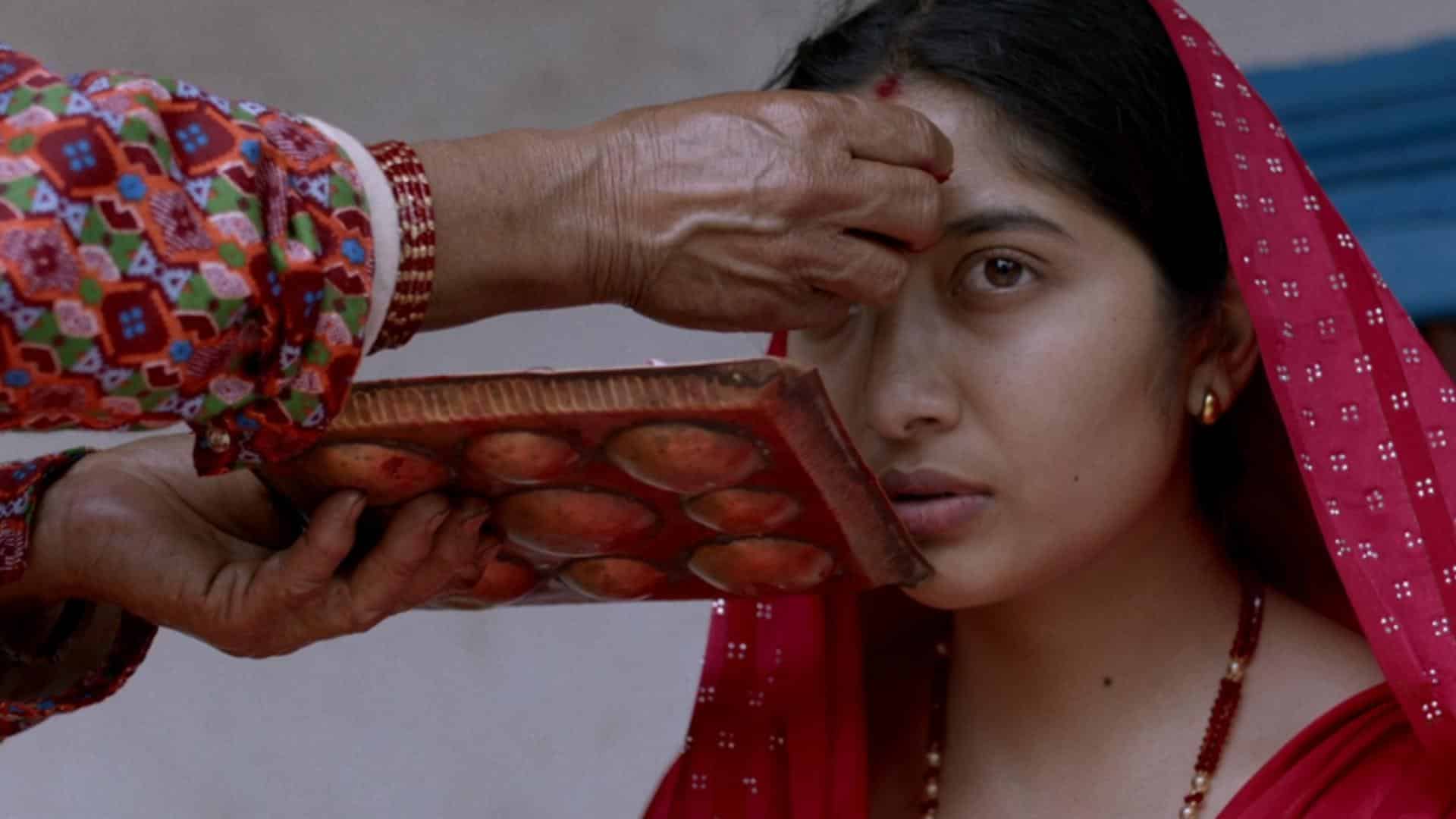 A film still from Eternal Kinship shows a Nepali woman with a soft gaze, looking at something off screen. She is wearing a red scarf draped over the crown of her head with a red top and a necklace with red beads and pearls. Coming into frame from the left are a pair of hands. One hand touches the forehead of the woman while the other hand holds a small gold platter.