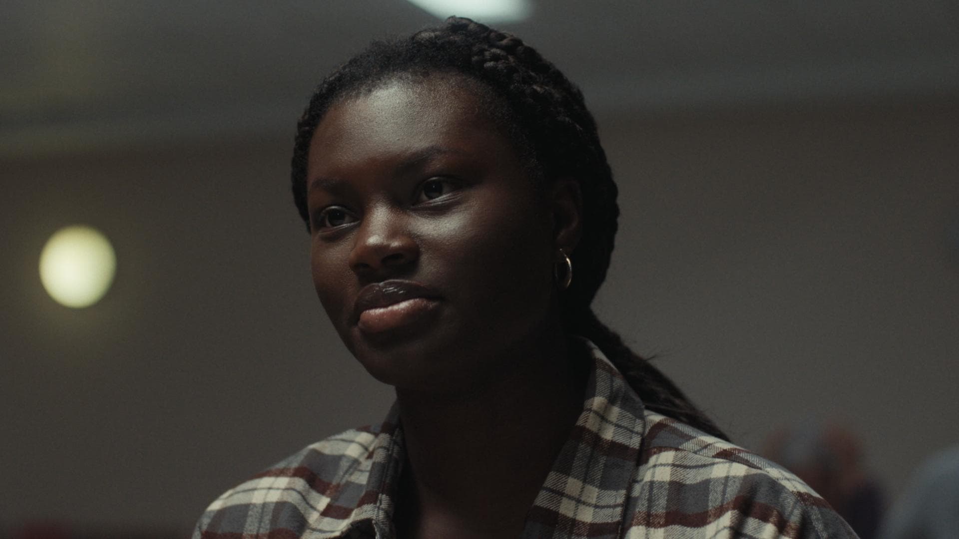 A film still from Lana shows a young Black woman with a calm gaze fixed off screen. She wears a gray, white, and red flannel with her hair in braids.