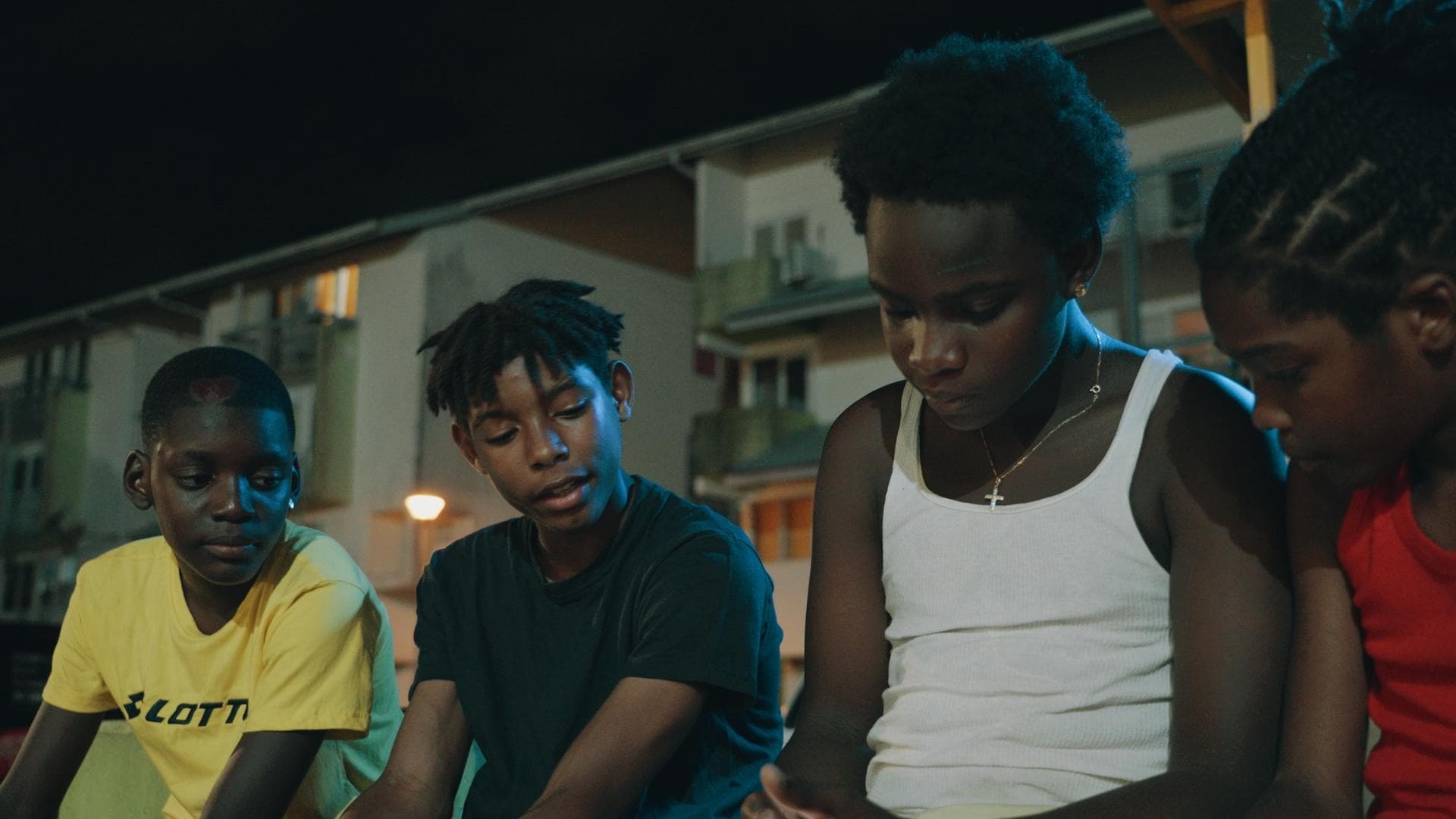 A film still from Listen to the Voices shows four young Black boys sitting side-by-side. Their attention is focused on something that one of the boys holds in his lap off screen.