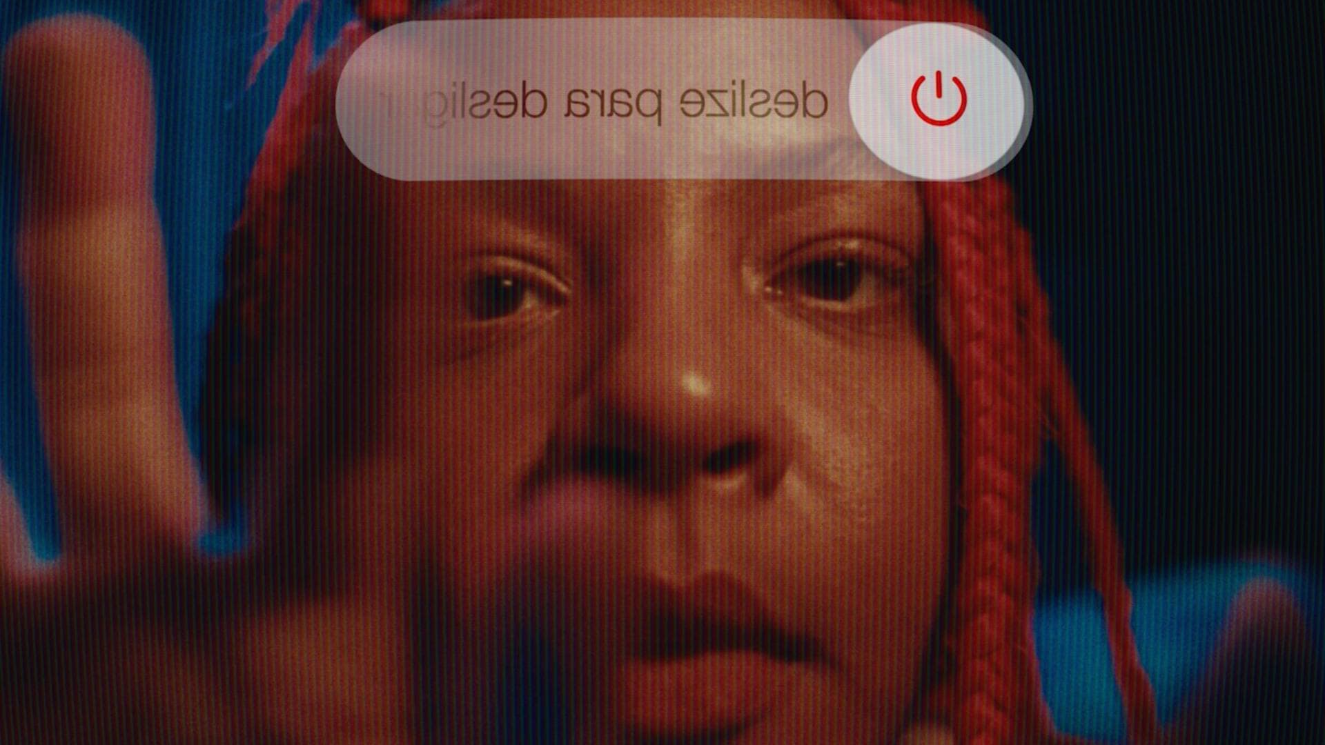A film still from Lazy White Cows shows the face of a Brazilian girl with red braids from the perspective of her phone. The power off symbol can be seen at the top of the still. Her hand can be seen reaching to turn off her phone.