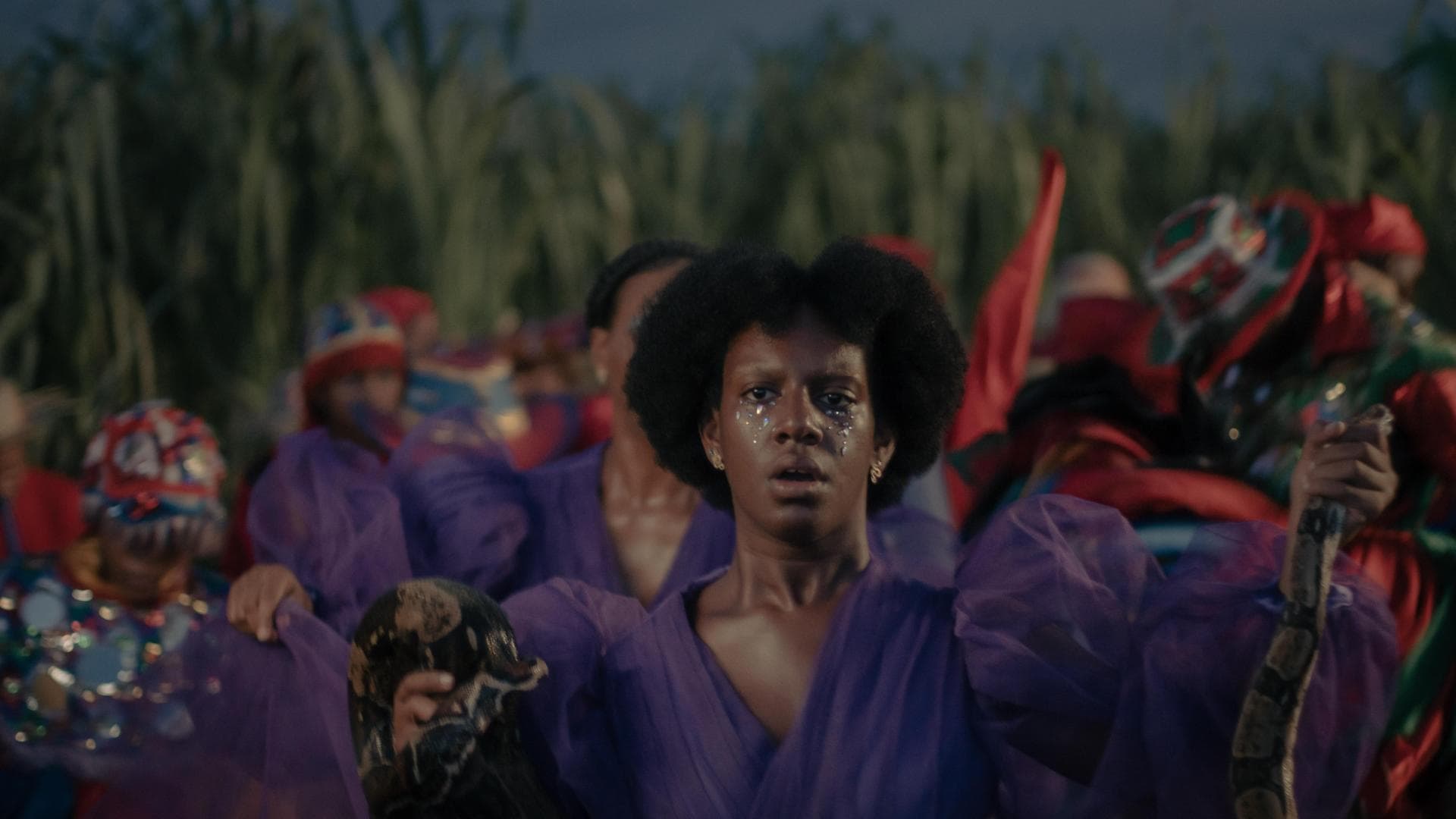 A film still from Sugar Island shows a young Dominican-Haitian woman in a purple tulle dress holding a snake between her hands. Her mouth is slightly open and her eyes, adorned with small gems beneath them are focused with concern on something beyond the camera. In the blurred background, more people can be seen wearing similar purple dresses, along with others in red and green outfits.