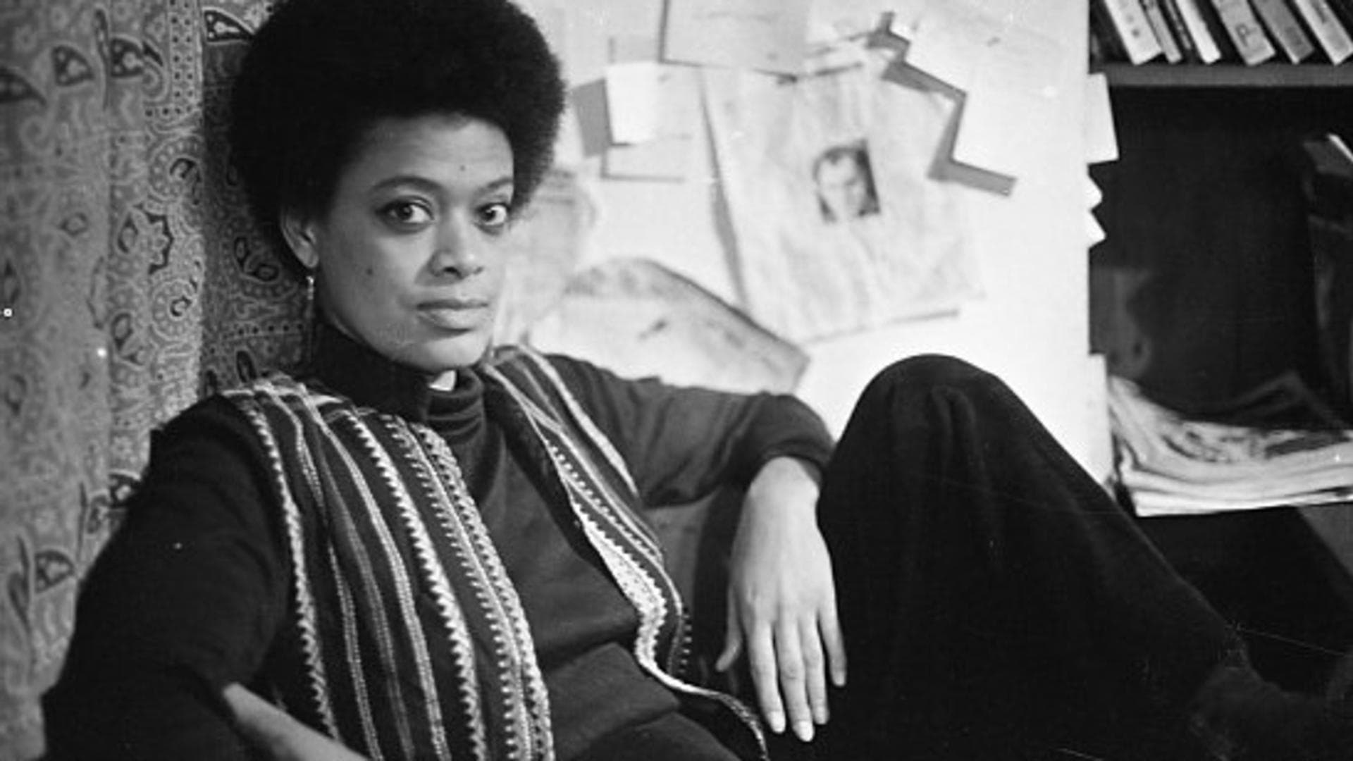 A black-and-white film still from TCB – The Toni Cade Bambara School of Organizing shows Toni Cade Bambara leaning against a paisley-printed curtain, gazing directly at the camera. She wears an afro and an open striped vest over a long-sleeve top. Behind her is a board with papers pinned to it and a shelf with books.