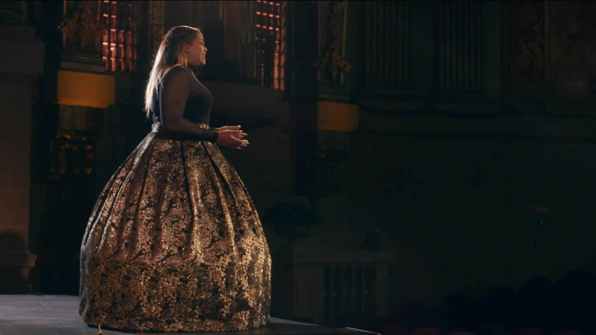 A film still from Tessitura shows a Black woman standing on the stage of a grand room, presumably singing. Her elbows are bent with hands in front of her. She wears a black sheer long-sleeved top and a black floor length skirt with a gold pattern.