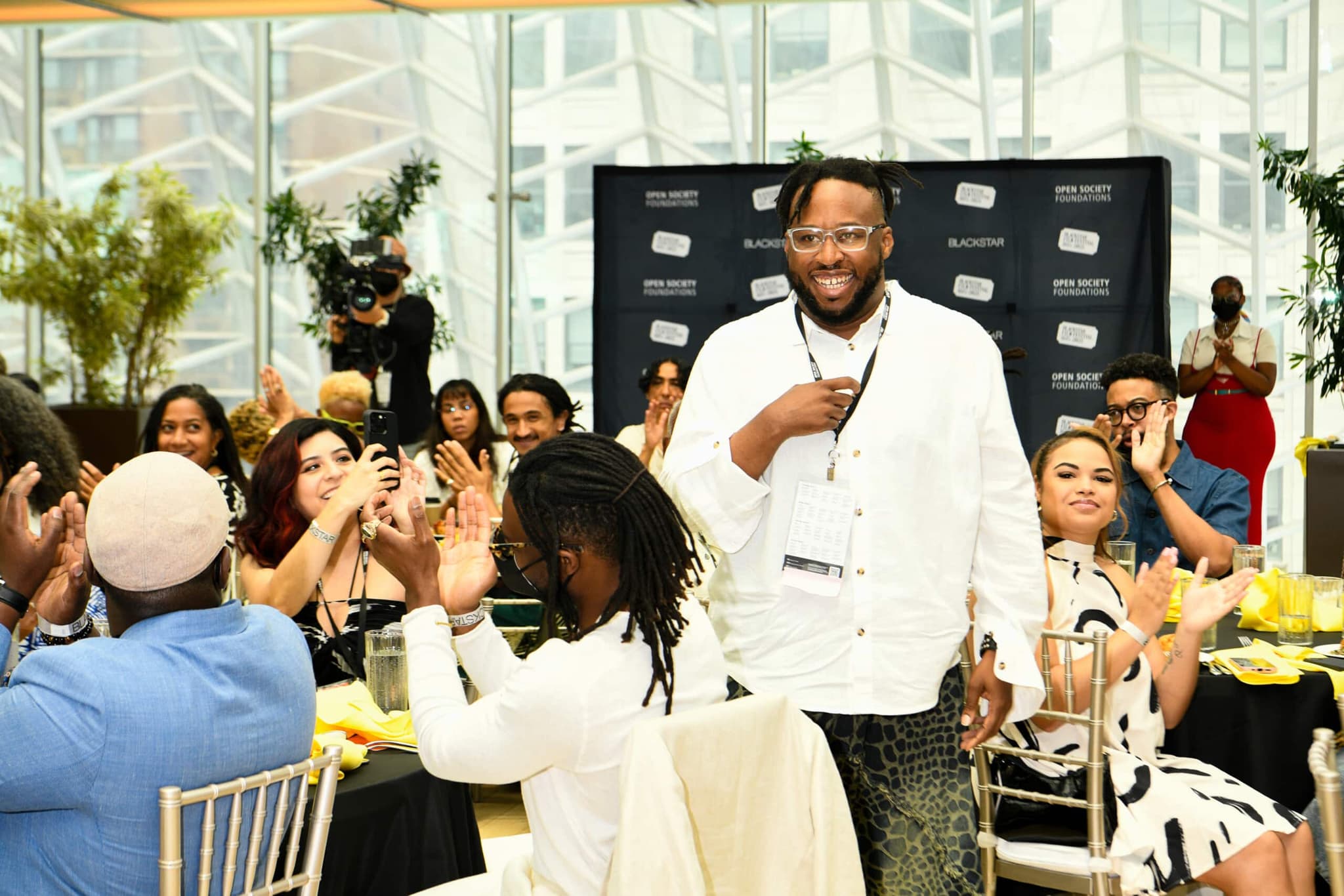 A photo from the Director's Brunch at BlackStar Film Festival. There is someone in the middle walking and smiling, others around them are seated in tables and applauding.