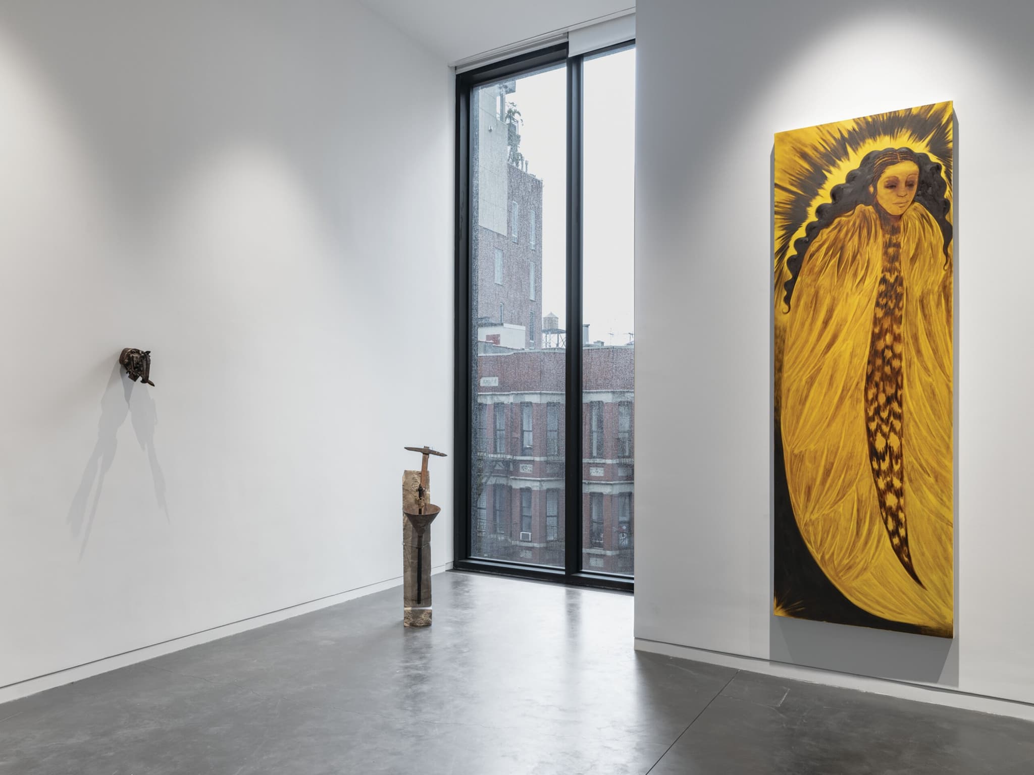 An artwork of a woman with wings hung up on a wall next to a wall-length window. The painting is mainly yellow gold with a little bit of black for contrast.