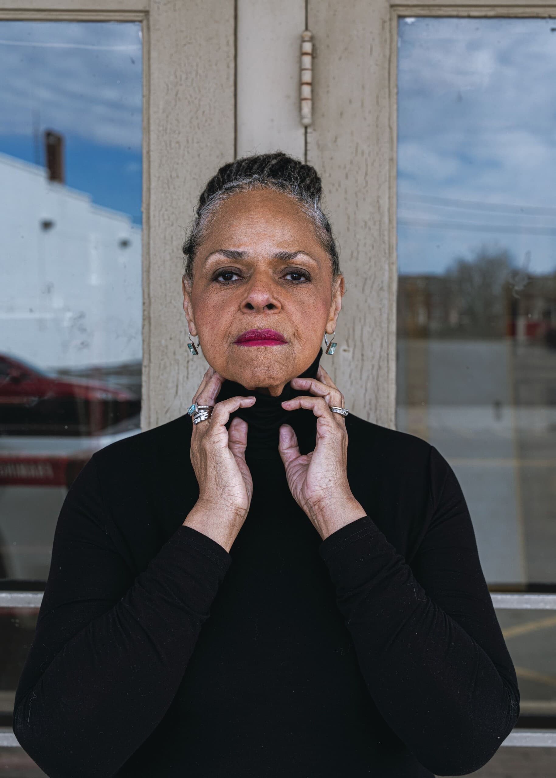 Beverly Wood posed with her hands by her neck in front of a door, looking directly at the camera.