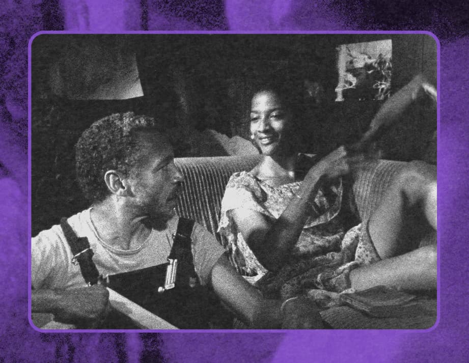Still from Losing Ground, a woman is sitting on the couch with her legs folded, and a man in overalls is sitting down beside her on the floor.