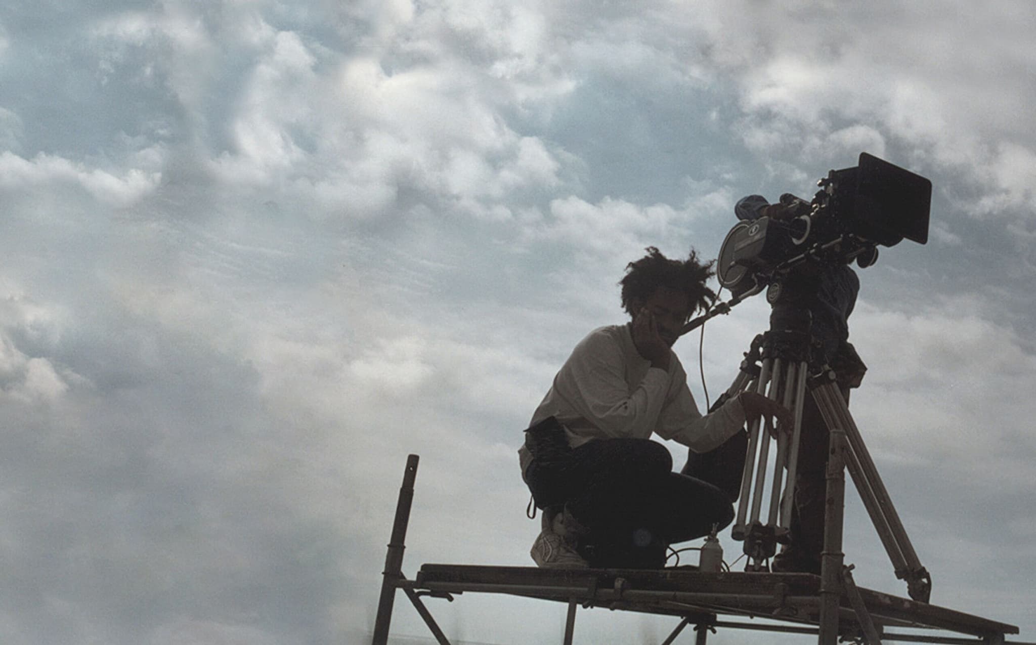 A picture of Arthur Jafa on the film set of Daughters of the Dust with the camera equipment on the right side. He is kneeling on some kind of elevated surface.