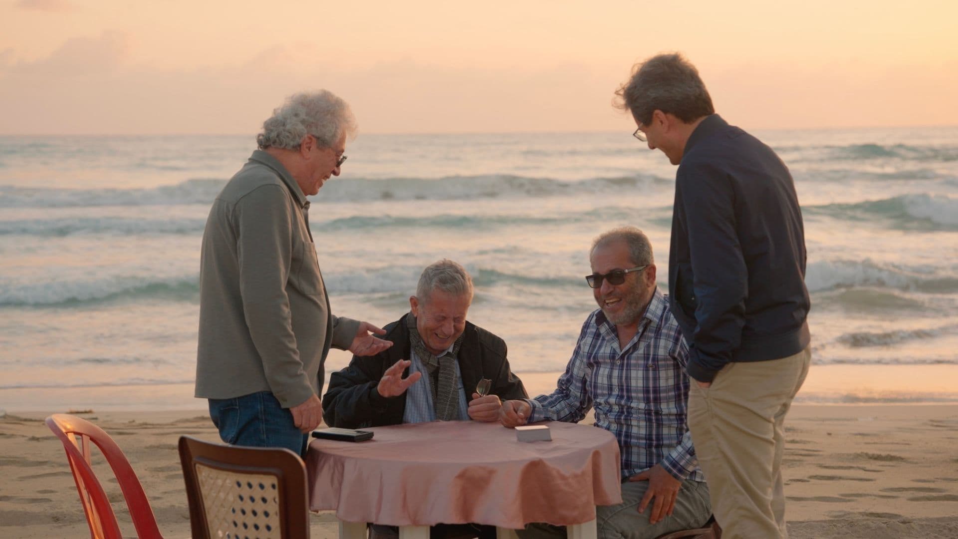 A film still from 66 Days shows four older men laughing around a round table on a sandy beach. The two men in the center are seated while the men on the left and right side are standing and looking down at them. In the background, the waves are crashing on the beach and the sky is a soft pink
