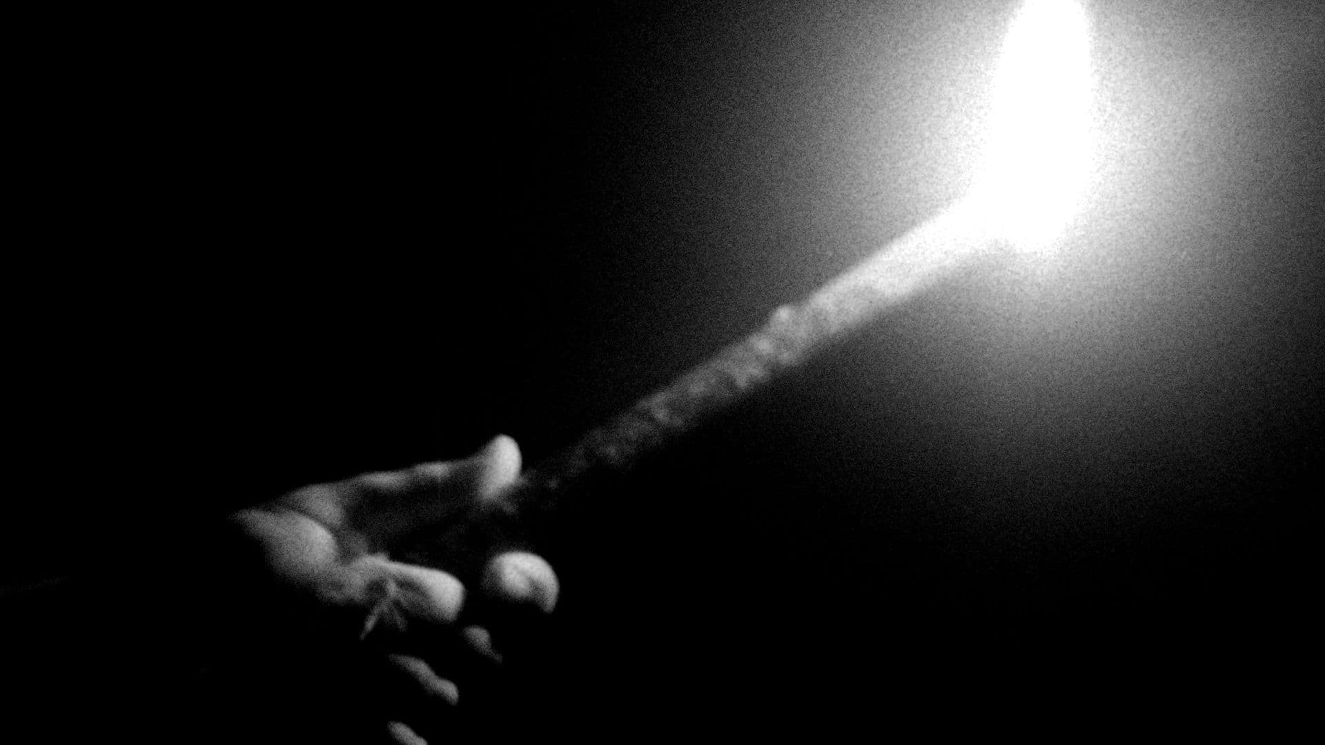 A black-and-white film still from A Bundle of Silences shows a hand holding a lit flare. The flare is releasing smoke and barely illuminates the hand.