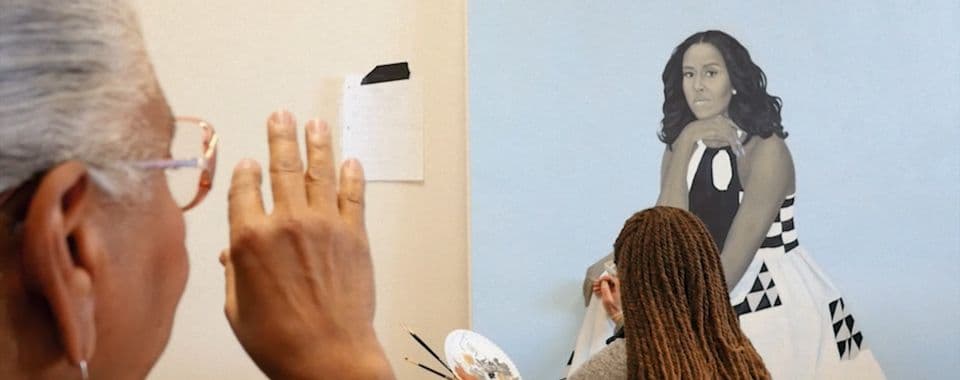 A film still from A Life in Art: Through the Eye of Dr. Leslie King Hammond shows Hammond facing away from the frame, gesturing as she watches a young Black painter who is adding finishing touches on a portrait of Michelle Obama. The painter is holding a brush and palette in her left hand and painting Obama’s hand.