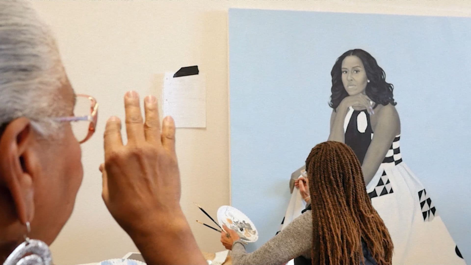 A film still from A Life in Art: Through the Eye of Dr. Leslie King Hammond shows Hammond facing away from the frame, gesturing as she watches a young Black painter who is adding finishing touches on a portrait of Michelle Obama. The painter is holding a brush and palette in her left hand and painting Obama’s hand.