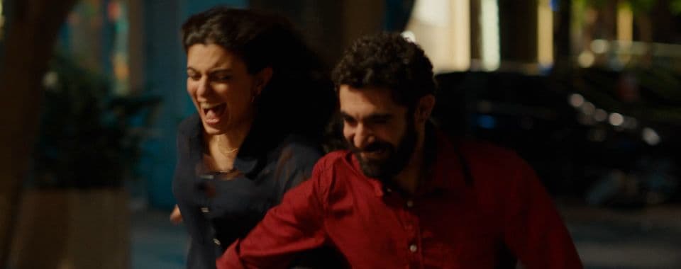 A film still from A Sad and Beautiful World shows a smiling Lebanese man and a laughing Lebanese woman running on a street at night. The man is wearing a red shirt and the woman is wearing a blue dress.
