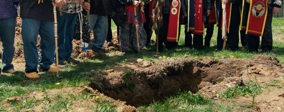 A film still from Aanikoobijigan (ancestor/great-grandparent/great-grandchild) shows a group of Native people standing around a freshly dug rectangular pit on a sunny day. The frame is centered on the ground, showing the grass, the pit, and the lower bodies of the people.