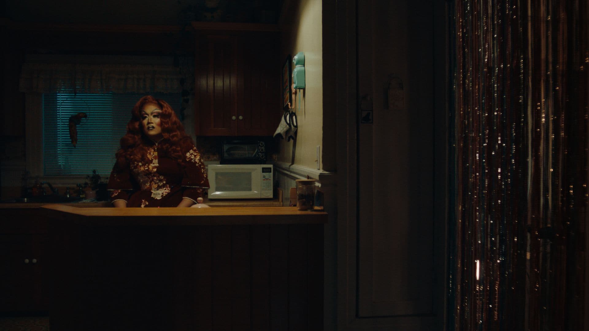 A film still from After What Happened at the Library shows an Asian drag queen sitting in a kitchen, staring into the distance. She is sitting behind a counter, wearing a black dress with red and white floral patterns and a shoulder-length red wavy wig. The kitchen is lit by a single light that throws harsh shadows across the room.