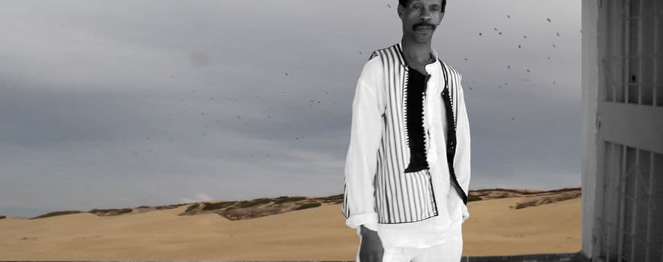 A film still from An Afternoon with a Gnawa shows a black-and-white image of a Black man standing in front of a view of the desert. The man is facing forward, wearing a striped vest and a square cap. In the distance, there’s a flock of birds across a gray sky.
