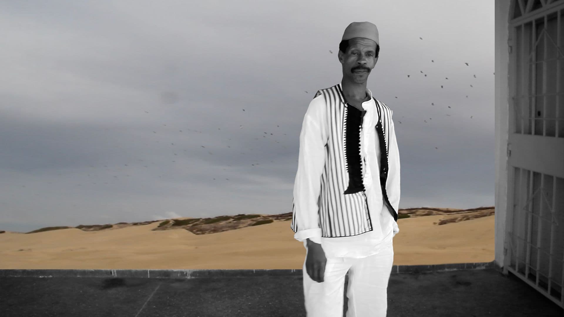 A film still from An Afternoon with a Gnawa shows a black-and-white image of a Black man standing in front of a view of the desert. The man is facing forward, wearing a striped vest and a square cap. In the distance, there’s a flock of birds across a gray sky.