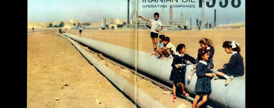 A scanned image from An Incomplete Calendar shows a group of Iranian children playing on a pipeline in the desert. A few boys stand and balance on the pipeline while two young girls sit on top and talk to two other girls standing in the sand. In the background, there’s an oil refinery. In the upper right corner of the image, it reads: “Iranian Oil Operating Companies 1958.”