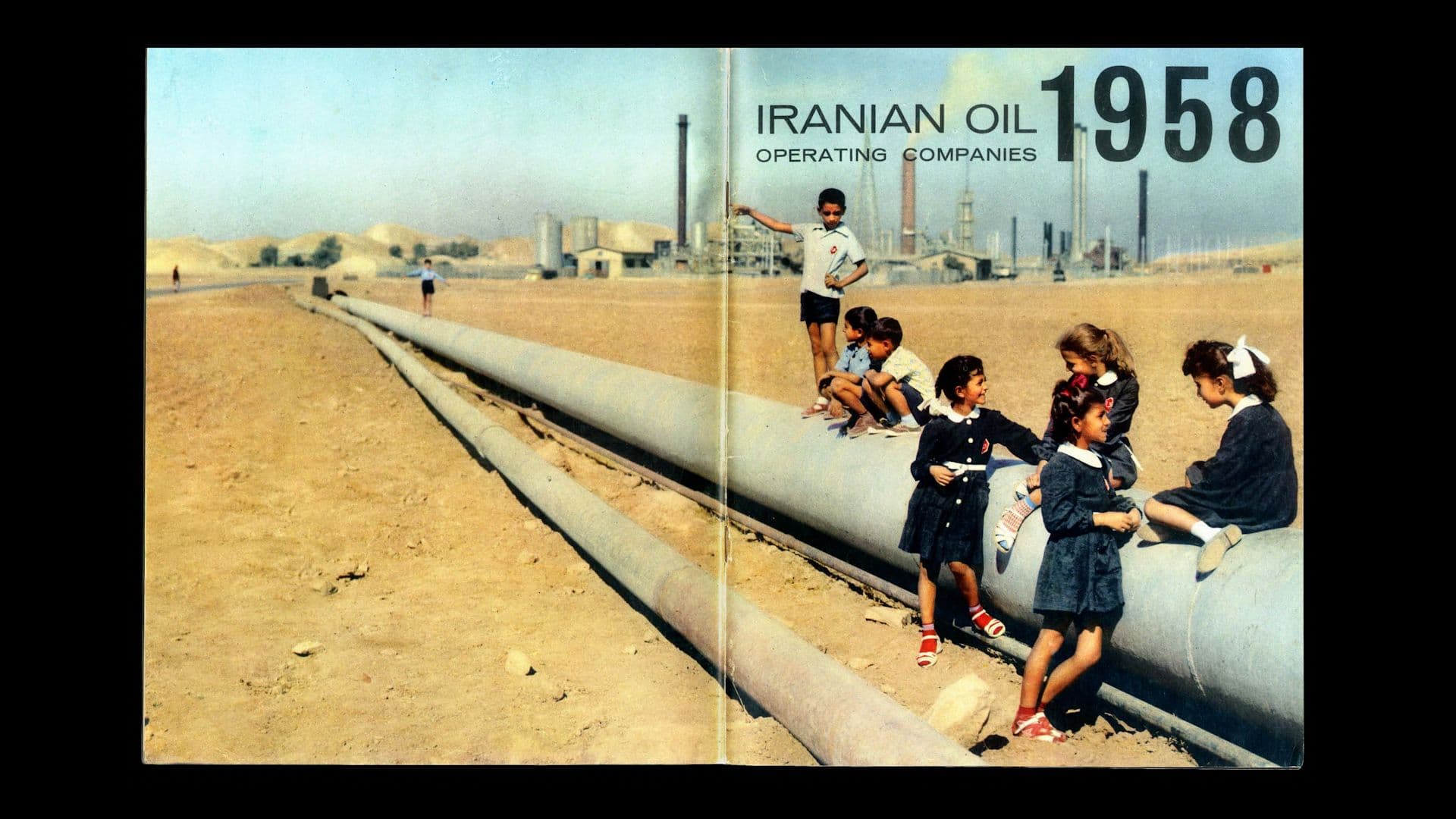 A scanned image from An Incomplete Calendar shows a group of Iranian children playing on a pipeline in the desert. A few boys stand and balance on the pipeline while two young girls sit on top and talk to two other girls standing in the sand. In the background, there’s an oil refinery. In the upper right corner of the image, it reads: “Iranian Oil Operating Companies 1958.”