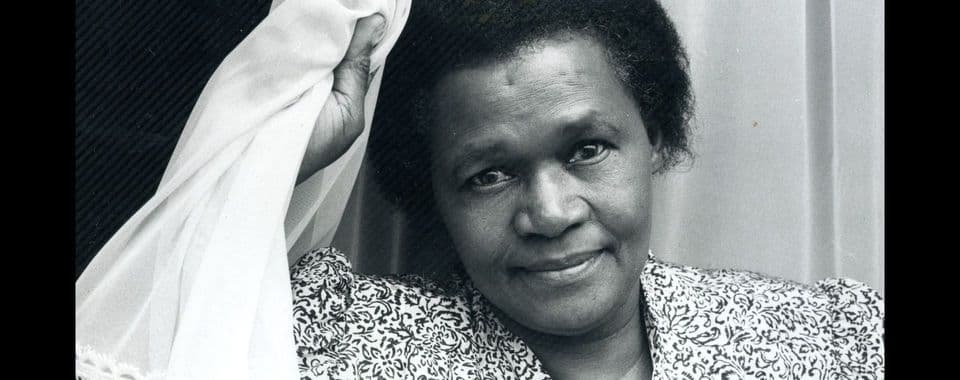 A black-and-white film still from And She Didn’t Die shows a Black woman facing the camera with a soft smile. She’s wearing a patterned shirt and holding a white curtain near her face using her right hand.