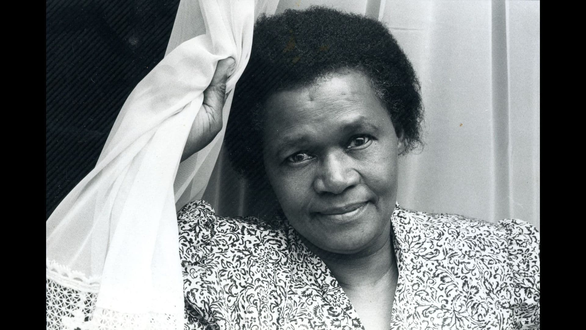 A black-and-white film still from And She Didn’t Die shows a Black woman facing the camera with a soft smile. She’s wearing a patterned shirt and holding a white curtain near her face using her right hand.