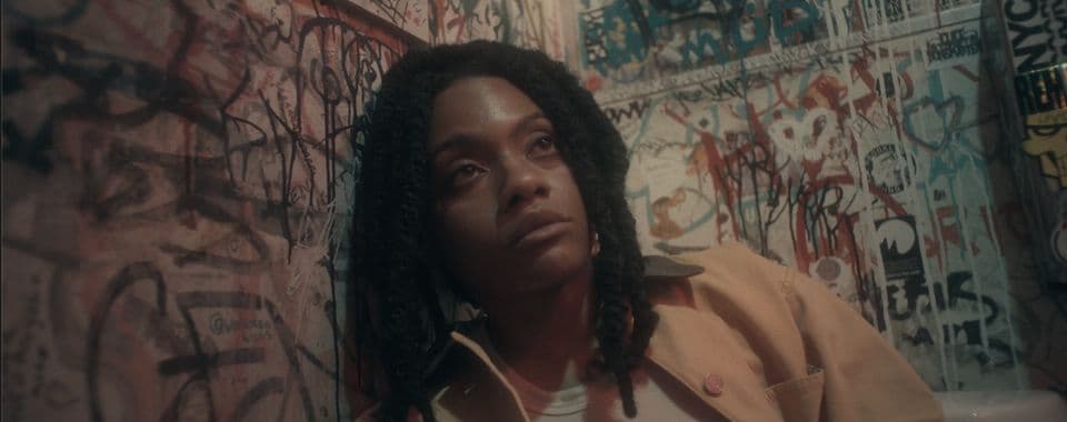 A film still from Angels Pull Your Hair shows a young Black woman leaning against a graffiti-covered wall in a bathroom. She looks upward with a solemn expression.