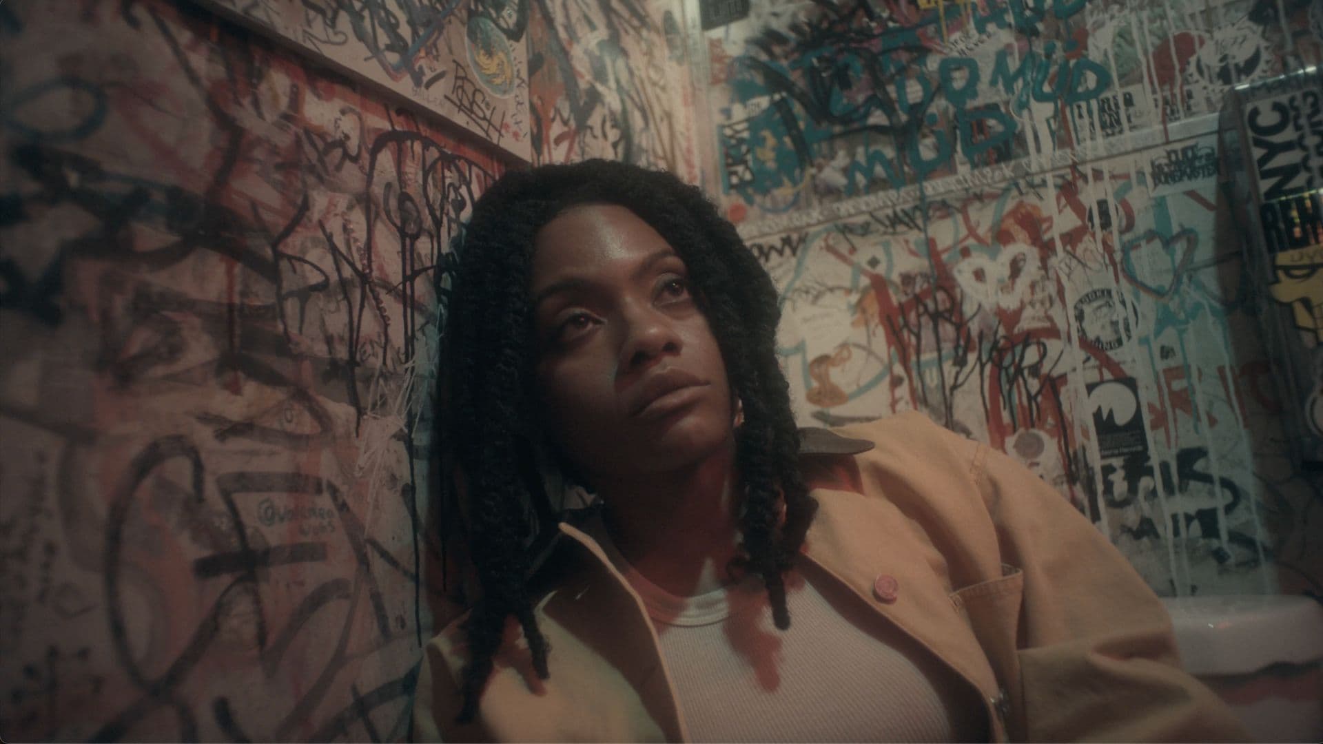 A film still from Angels Pull Your Hair shows a young Black woman leaning against a graffiti-covered wall in a bathroom. She looks upward with a solemn expression.