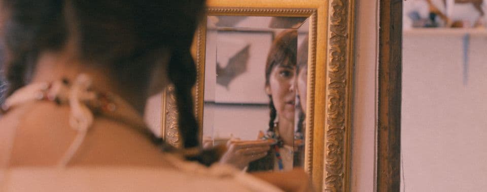 A film still from Are You Native? shows a woman standing in front of a mirror looking at herself. The mirror reveals half of her face and her left hand softly touching her shoulder. Her hair is braided and she is wearing traditional regalia.