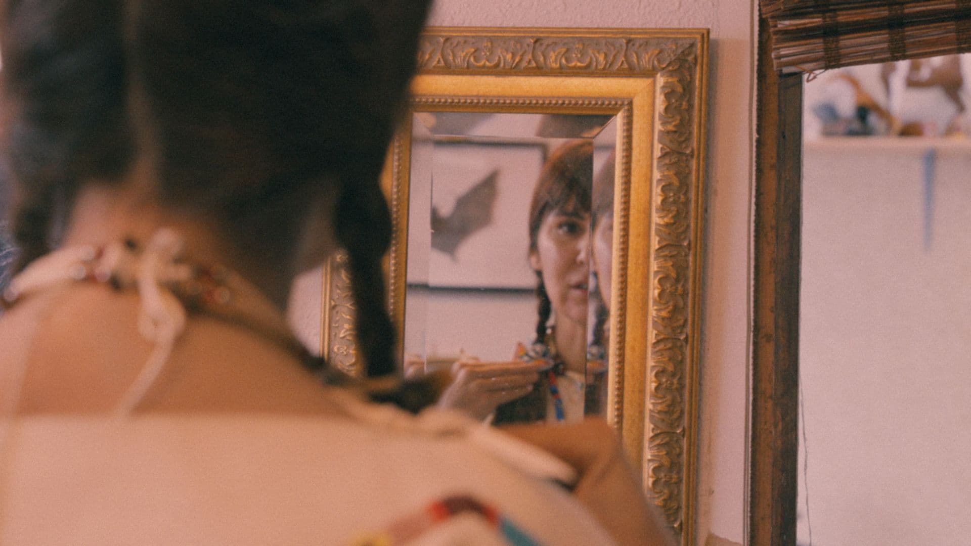 A film still from Are You Native? shows a woman standing in front of a mirror looking at herself. The mirror reveals half of her face and her left hand softly touching her shoulder. Her hair is braided and she is wearing traditional regalia.
