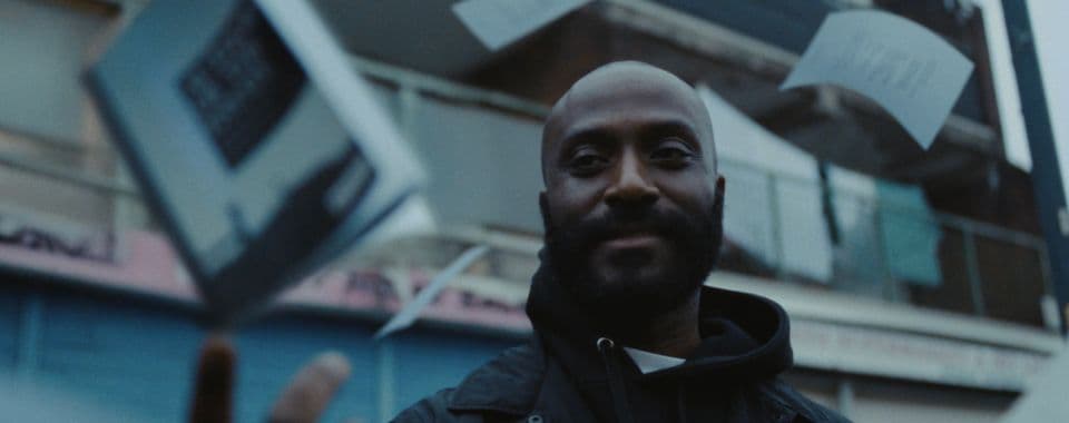 A film still from Authors of the Estate shows a smiling Black man in a black jacket looking towards a paperback book that’s mid-air above his right hand. Loose pages fly around him as he stands in front of a worn apartment building.