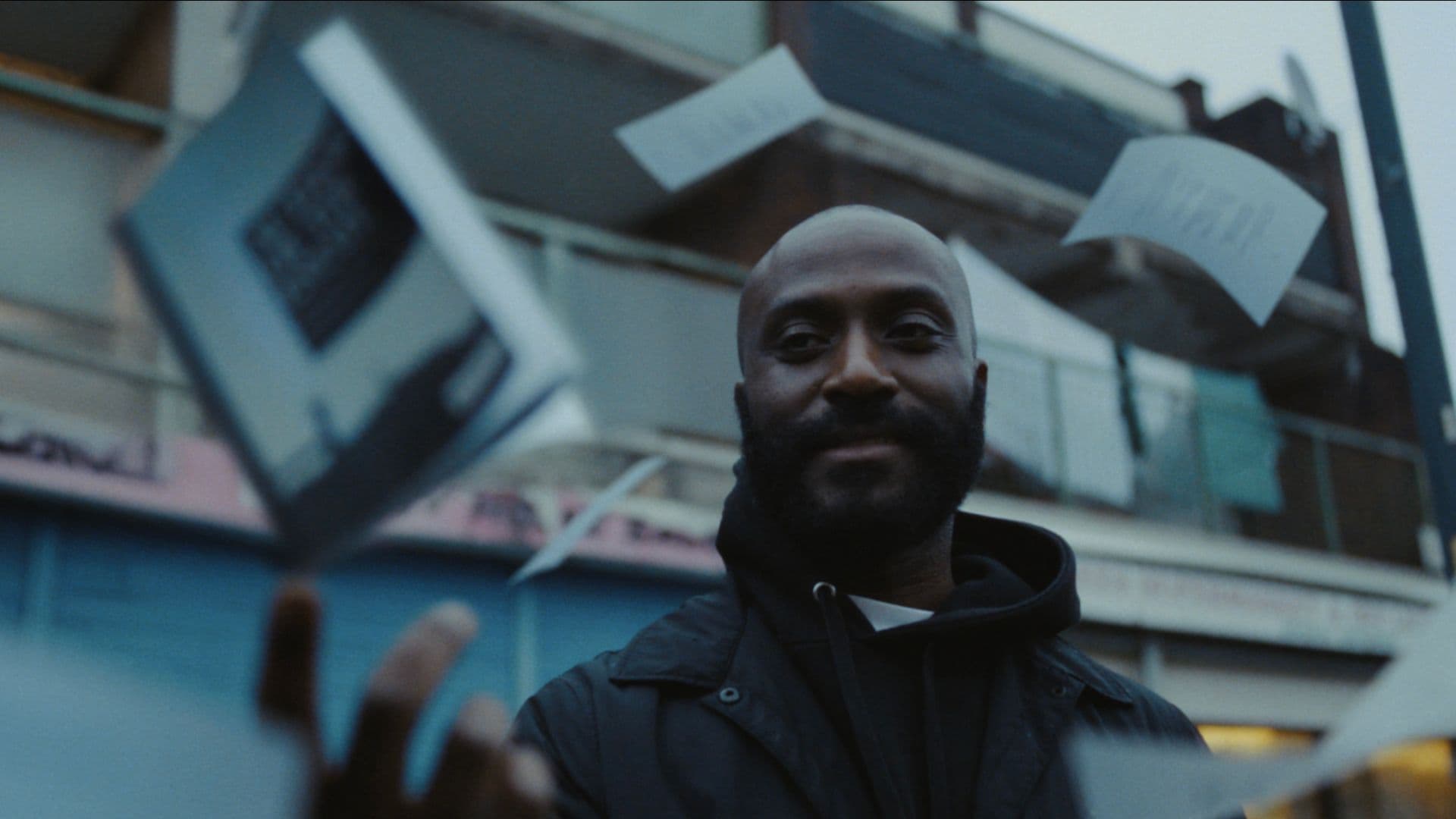 A film still from Authors of the Estate shows a smiling Black man in a black jacket looking towards a paperback book that’s mid-air above his right hand. Loose pages fly around him as he stands in front of a worn apartment building.