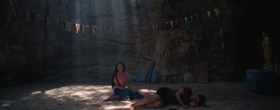 A film still from Becoming Human shows a seated teenage girl and sleeping woman on a tiled floor in a cave. The girl is wearing a pink shirt and blue skirt and is staring off to the left. The woman is lying on her left side. There’s a line of multi-colored flags stretching across the cave wall and a gold statue on the right.