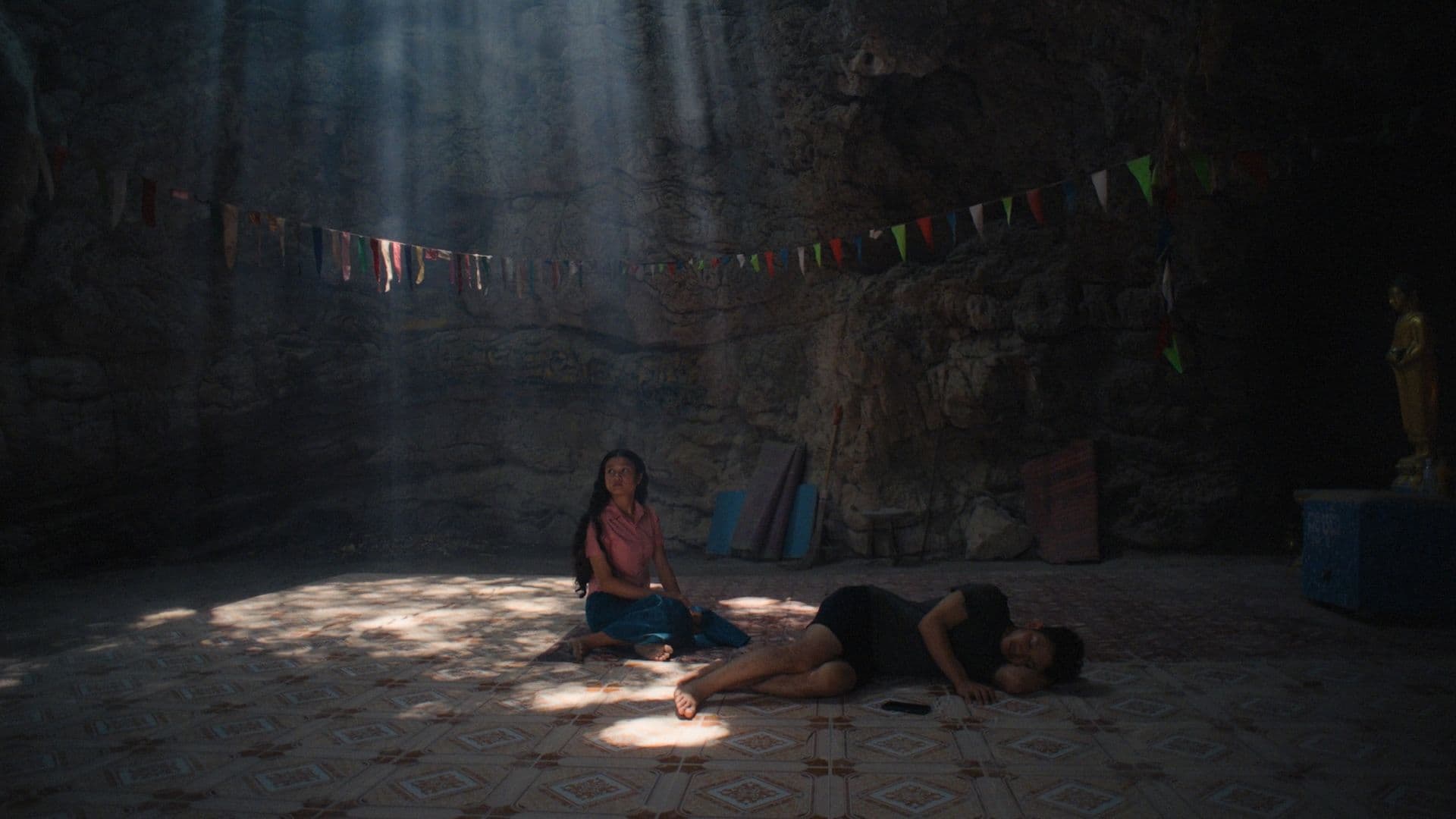 A film still from Becoming Human shows a seated teenage girl and sleeping woman on a tiled floor in a cave. The girl is wearing a pink shirt and blue skirt and is staring off to the left. The woman is lying on her left side. There’s a line of multi-colored flags stretching across the cave wall and a gold statue on the right.