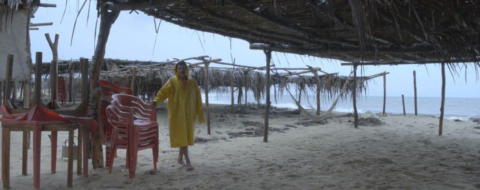 A film still from Boca Vieja shows a middle-aged man in a yellow rain jacket standing on a beach below a thatched roof. He holds onto a stack of pink stackable chairs.