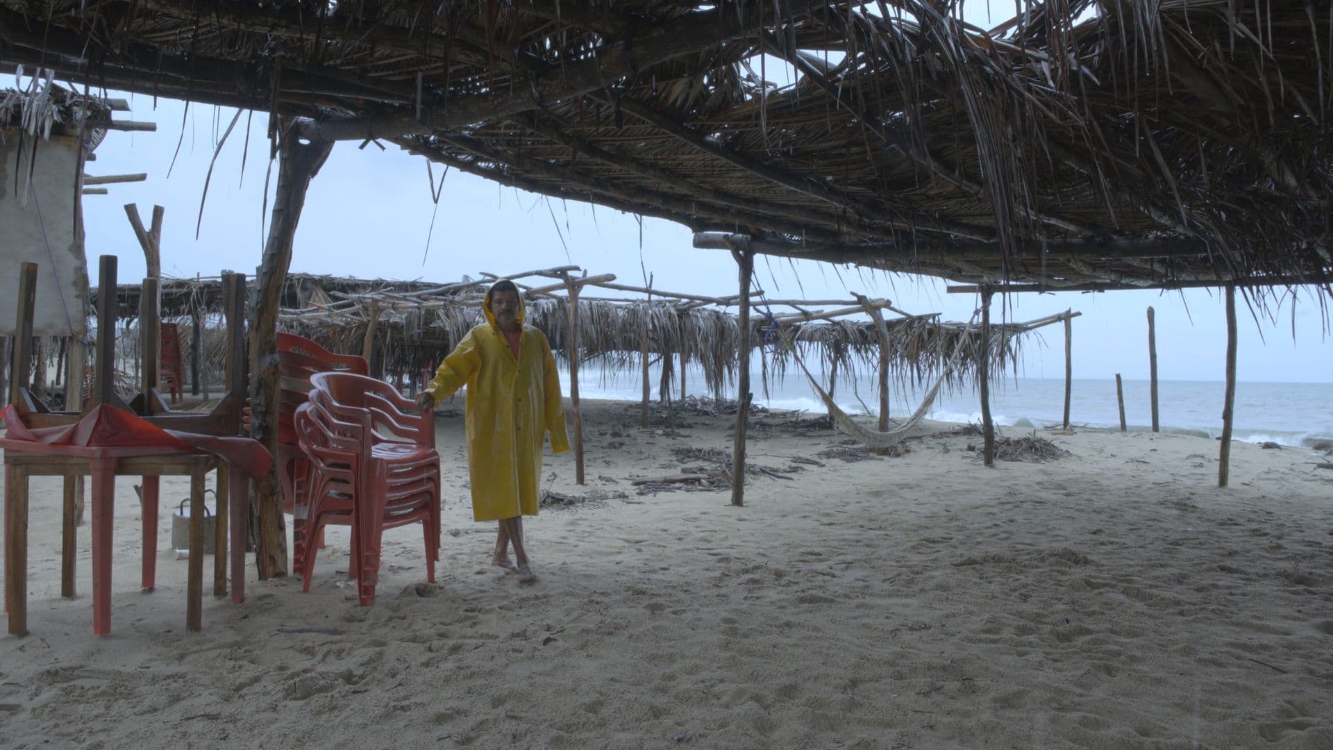 A film still from Boca Vieja shows a middle-aged man in a yellow rain jacket standing on a beach below a thatched roof. He holds onto a stack of pink stackable chairs.