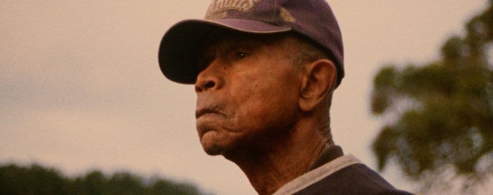 A film still from Buckskin shows an older Black man wearing a purple baseball cap looking off into the distance.